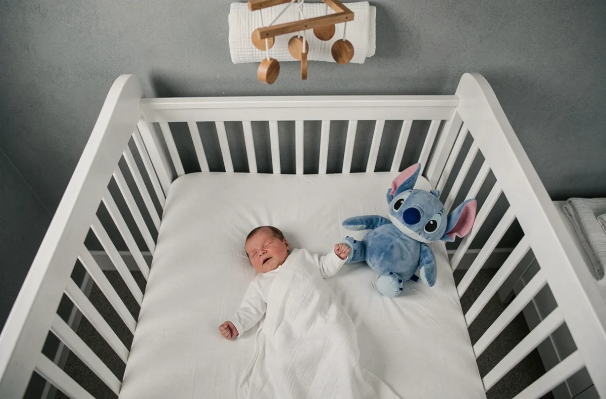 Nouveau-né dans un berceau en bois blanc avec une peluche Stitch posée à proximité en sécurité